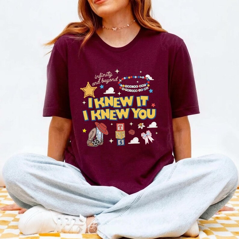 May include: A maroon short-sleeve t-shirt featuring a graphic print with the text "infinity and beyond", "YOU'VE GOT A FRIEND IN ME", "I KNEW IT", and "I KNEW YOU". The design includes a yellow star, a beaded bracelet, a cowboy boot, and alphabet blocks.