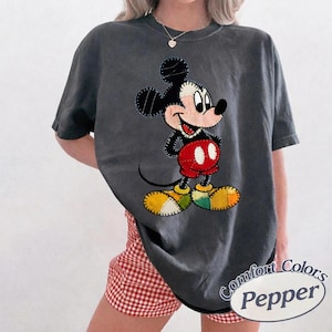 May include: Dark grey t-shirt with a Mickey Mouse graphic. The cartoon character has black ears, a white face, red shorts, and yellow shoes. The shirt has the words "Comfort Colors Pepper" printed on it.