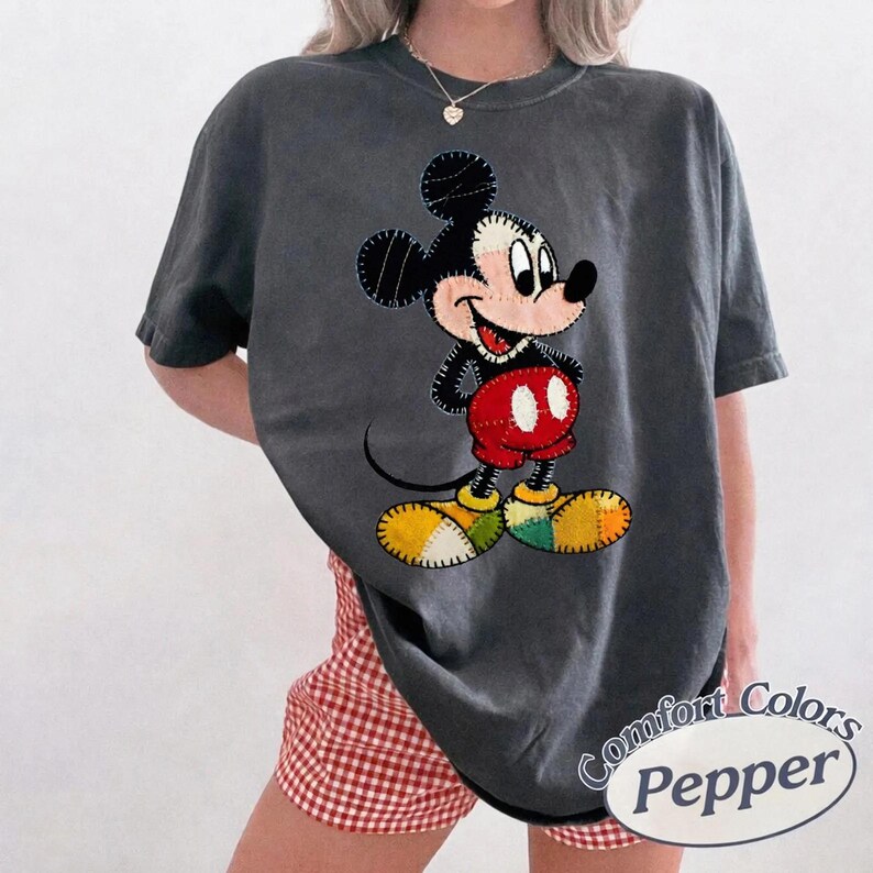 May include: Dark grey t-shirt with a Mickey Mouse graphic. The cartoon character has black ears, a white face, red shorts, and yellow shoes. The shirt has the words "Comfort Colors Pepper" printed on it.