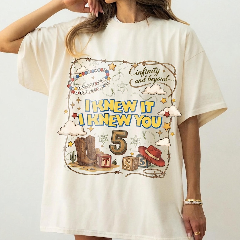 May include: An off-white t-shirt featuring a graphic print with a lasso border, cowboy boots, a red hat, alphabet blocks, and the text "Cinfinity and beyond", "I KNEW IT I KNEW YOU", "5", and "YOU'VE GOT A FRIEND IN ME" with stars and clouds.