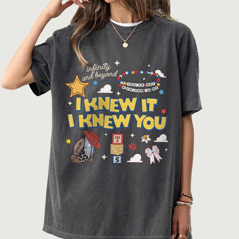 May include: A charcoal gray short-sleeve t-shirt featuring a graphic print with the text "infinity and beyond", "YOU'VE GOT A FRIEND IN ME", "I KNEW IT", and "I KNEW YOU". The design includes a yellow star, a beaded bracelet, a cowboy boot, blocks, and clouds.