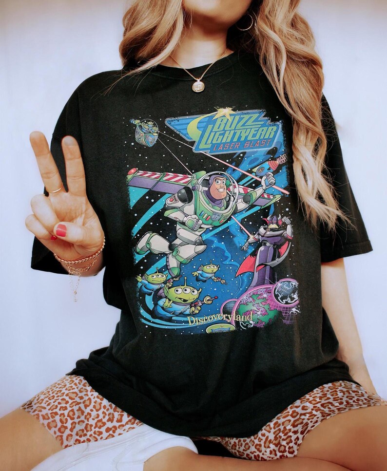 May include: A black t-shirt featuring a graphic print of Buzz Lightyear, Zurg, and green aliens in space. The text reads Buzz Lightyear Laser Blast and Discoveryland. The design includes colorful stars, planets, and laser beams on a dark background.