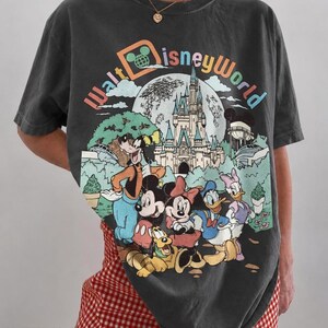 May include: Dark gray t-shirt featuring a colorful Walt Disney World graphic with Mickey Mouse, Minnie Mouse, Donald Duck, Goofy, and Pluto. The shirt has a vintage look and the text "COMFORT COLORS PEPPER" is at the bottom.