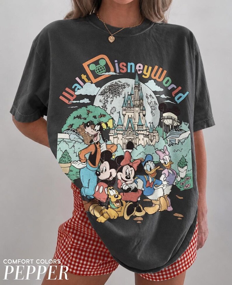 May include: Dark gray t-shirt featuring a colorful Walt Disney World graphic with Mickey Mouse, Minnie Mouse, Donald Duck, Goofy, and Pluto. The shirt has a vintage look and the text "COMFORT COLORS PEPPER" is at the bottom.