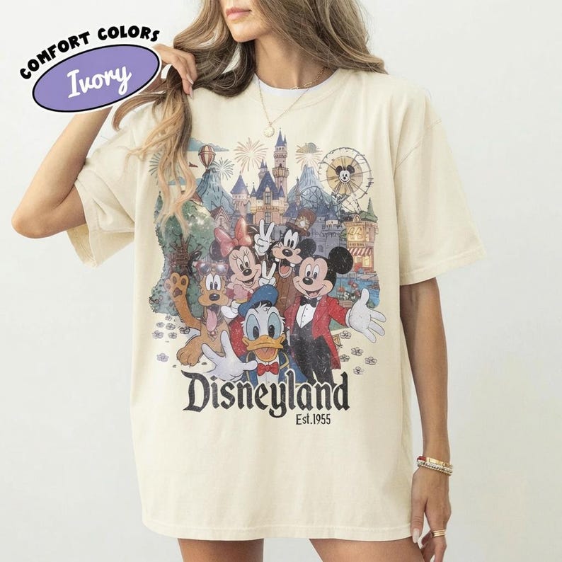 May include: An ivory short-sleeve t-shirt featuring a graphic print of Mickey Mouse, Minnie Mouse, Donald Duck, Goofy, and Pluto in front of a castle. The text reads Disneyland Est. 1955. A Comfort Colors logo is printed in the upper left corner.