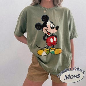 May include: An olive green t-shirt with a Mickey Mouse graphic. The cartoon character is wearing red shorts, yellow shoes, and black ears. The shirt has the words "Comfort Colors Moss" printed on it.