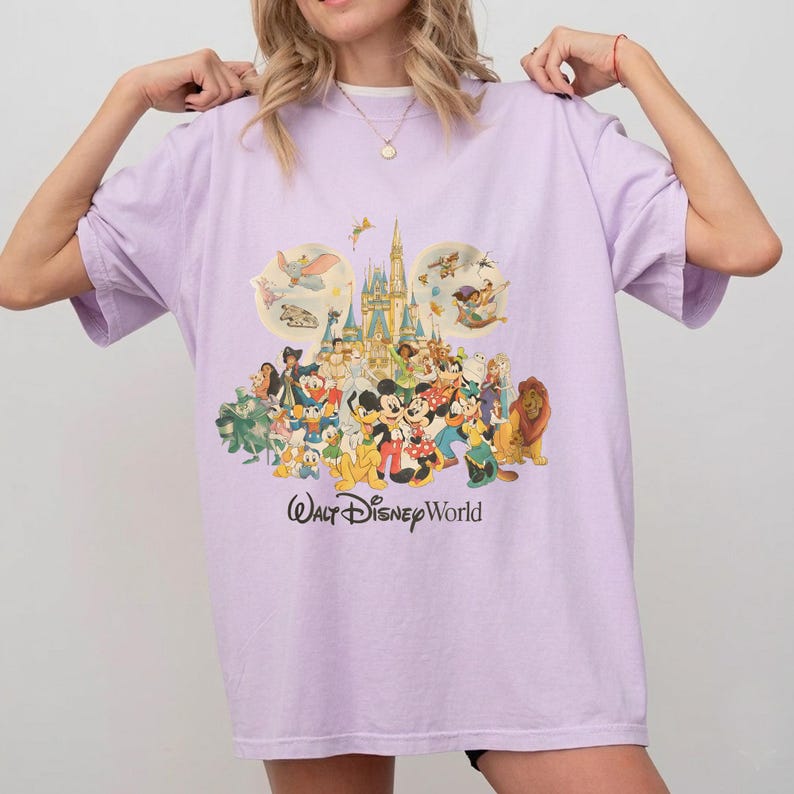 May include: A light purple t-shirt featuring a colorful graphic of Disney characters and a castle. The text "Walt Disney World" is printed in a script font below the graphic. The shirt has short sleeves and a crew neck.