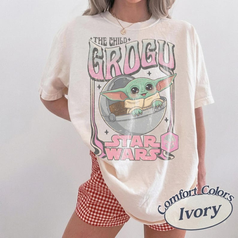 Puede incluir: Camiseta de color marfil con un gr&aacute;fico de Grogu de Star Wars. El dise&ntilde;o incluye el texto "THE CHILD + GROGU" en rosa, una imagen de Grogu en una c&aacute;psula y las palabras "STAR WARS" en rosa. La camiseta es de la marca "Comfort Colors Ivory".