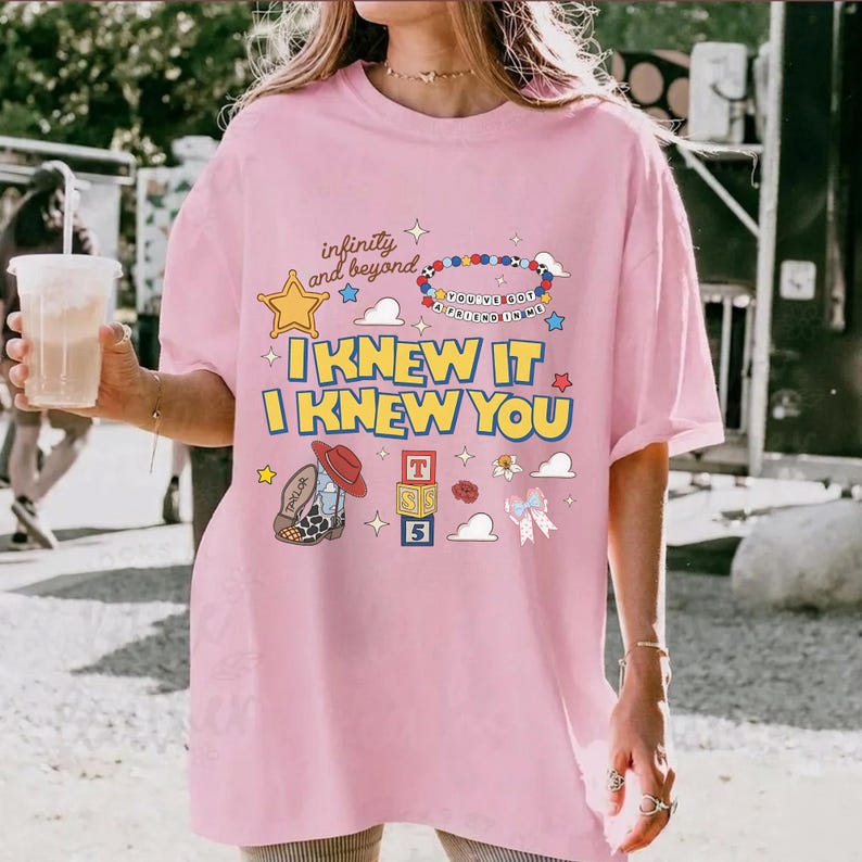 May include: A light pink t-shirt featuring a graphic print with the text "infinity and beyond", "YOU'VE GOT A FRIEND IN ME", "I KNEW IT", and "I KNEW YOU". The design includes a yellow star, a beaded necklace, a cowboy boot, alphabet blocks, and a bow.