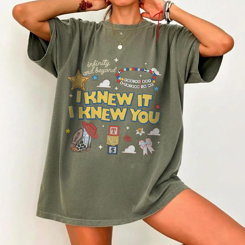 May include: An olive green short-sleeve t-shirt featuring a graphic print with the text "infinity and beyond," "YOU'VE GOT A FRIEND IN ME," and "I KNEW IT I KNEW YOU." The design includes a star, a beaded necklace, a cowboy boot, blocks, and a bow.