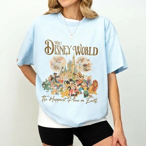 May include: Light blue t-shirt featuring the text "Walt Disney World" in gold script above a colorful graphic of Disney characters and the phrase "The Happiest Place on Earth."
