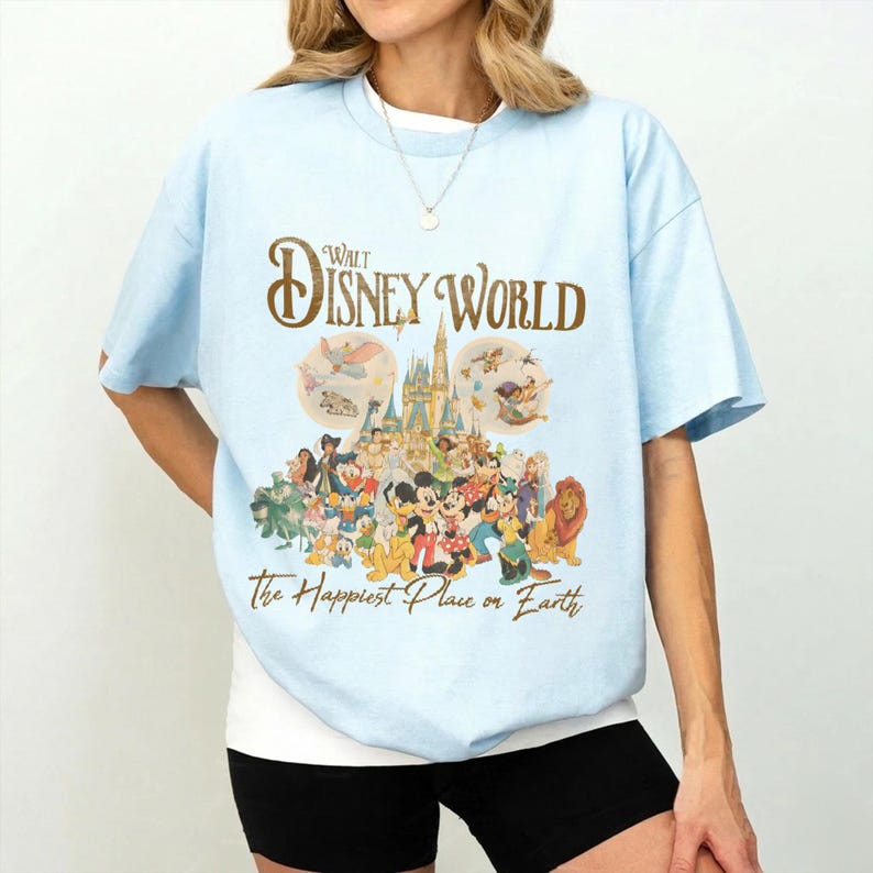 May include: Light blue t-shirt featuring the text "Walt Disney World" in gold script above a colorful graphic of Disney characters and the phrase "The Happiest Place on Earth."