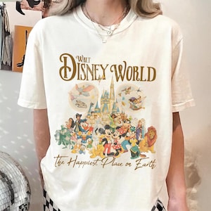May include: Cream-colored t-shirt featuring a colorful graphic of Disney characters, a castle, and the text "Walt Disney World" and "The Happiest Place on Earth" in brown. The design includes Mickey Mouse, Donald Duck, and other iconic characters.