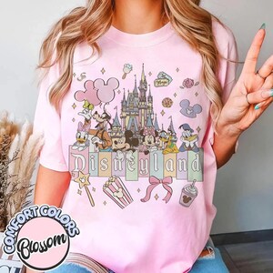 May include: Pink t-shirt featuring a Disneyland design. The graphic includes the Disneyland castle, Disney characters, and the word "Disneyland" in colorful letters. Additional elements include a popcorn box, bow, and Mickey Mouse-shaped balloons.