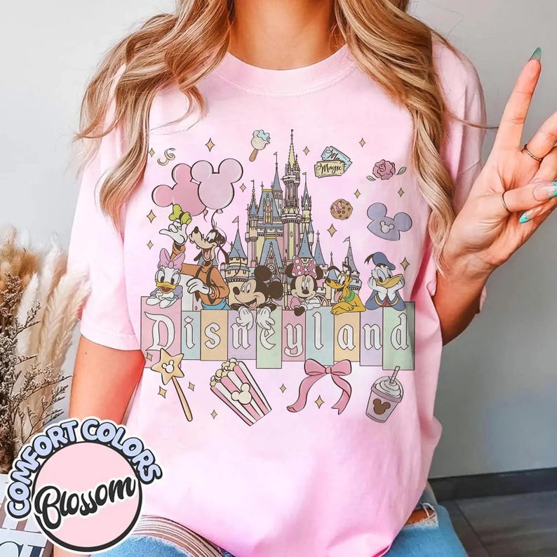 May include: Pink t-shirt featuring a Disneyland design. The graphic includes the Disneyland castle, Disney characters, and the word "Disneyland" in colorful letters. Additional elements include a popcorn box, bow, and Mickey Mouse-shaped balloons.