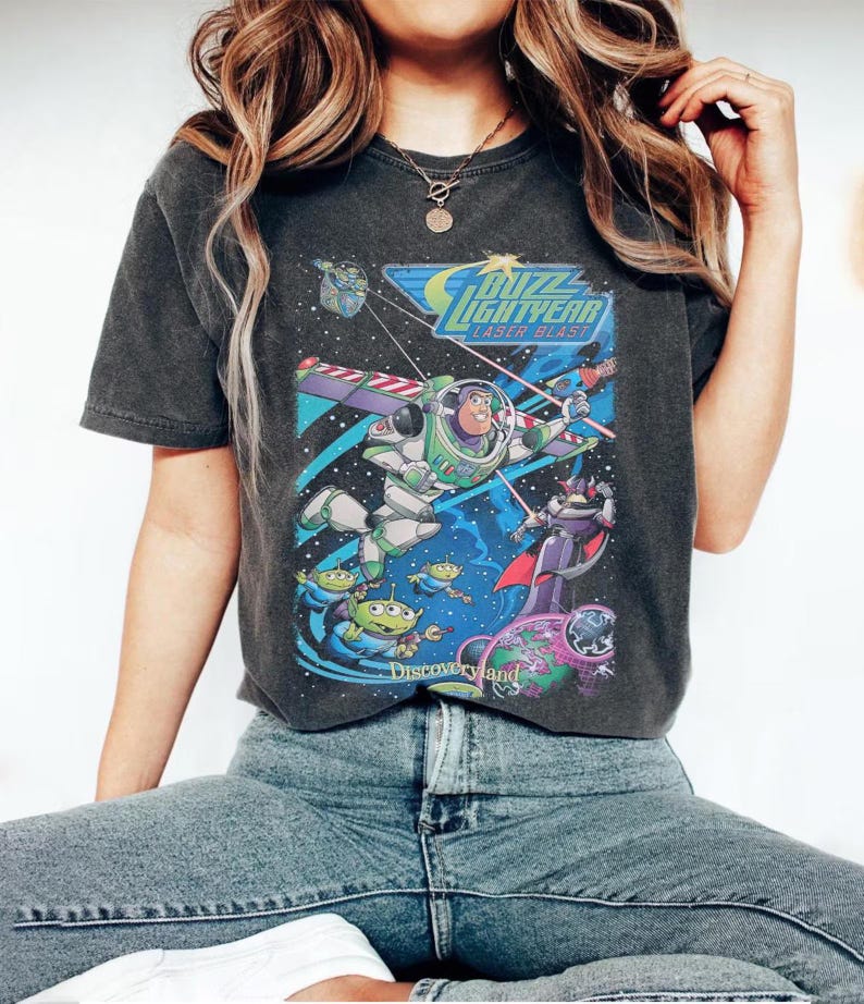 May include: A charcoal gray short-sleeve t-shirt featuring a graphic print of Buzz Lightyear Laser Blast. The design includes Buzz Lightyear, Zurg, and green aliens in space with the text Buzz Lightyear Laser Blast and Discoveryland.