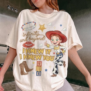 May include: A cream-colored t-shirt featuring a graphic of a cowgirl character, a red hat, brown cowboy boots, and alphabet blocks. The text reads "infinity and beyond" and "I KNEW IT I KNEW YOU" along with a beaded necklace graphic.