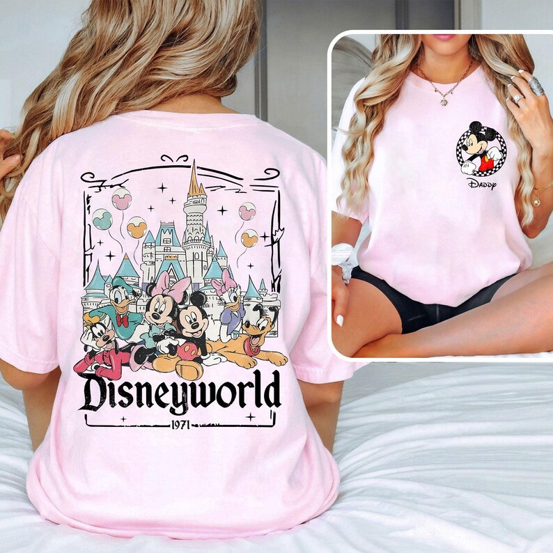 May include: A light pink t-shirt featuring a Disney World graphic on the back with Mickey Mouse, Minnie Mouse, Donald Duck, and other characters. The front of the shirt has a small Mickey Mouse graphic. The text "Disneyworld - 1971" is printed on the back.
