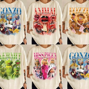 May include: A collection of six cream-colored t-shirts featuring graphic prints of Muppets characters including Gonzo, Animal, Fozzie Bear, Kermit the Frog, and Miss Piggy. Each shirt displays the character's name in large, bold, colorful lettering.