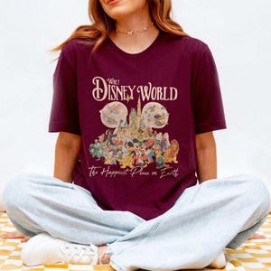 May include: Burgundy t-shirt featuring the text "Walt Disney World" above a graphic of Disney characters and the castle. The phrase "The Happiest Place on Earth" is printed below the graphic.