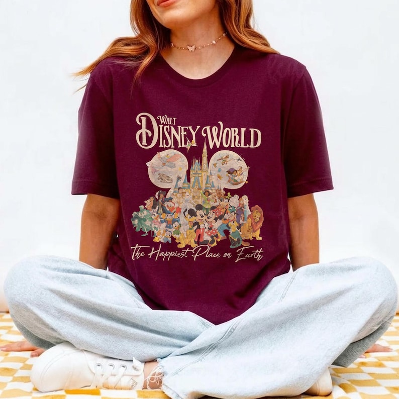 May include: Burgundy t-shirt featuring the text "Walt Disney World" above a graphic of Disney characters and the castle. The phrase "The Happiest Place on Earth" is printed below the graphic.