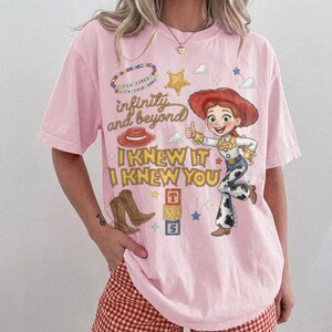 May include: A light pink short-sleeve t-shirt featuring a graphic print of a cowgirl character, a red hat, brown boots, and alphabet blocks. The text reads infinity and beyond, I KNEW IT, and I KNEW YOU, surrounded by stars and clouds.
