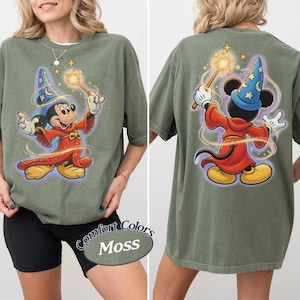 May include: A moss green t-shirt featuring a graphic of Mickey Mouse dressed as a wizard in a red robe and blue star-patterned hat, holding a glowing wand. The design appears on both the front and back of the shirt, with swirling light effects.