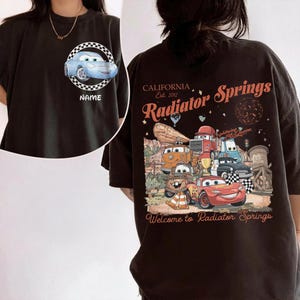 Puede incluir: Camiseta negra con un coche de dibujos animados azul claro en un c&iacute;rculo a cuadros y la palabra "NAME". La parte posterior de la camiseta muestra un gr&aacute;fico de varios coches de dibujos animados con el texto "CALIFORNIA Est. 2012 Radiator Springs" y "Welcome to Radiator Springs."