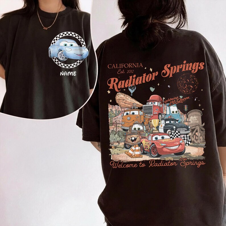 Puede incluir: Camiseta negra con un coche de dibujos animados azul claro en un c&iacute;rculo a cuadros y la palabra "NAME". La parte posterior de la camiseta muestra un gr&aacute;fico de varios coches de dibujos animados con el texto "CALIFORNIA Est. 2012 Radiator Springs" y "Welcome to Radiator Springs."