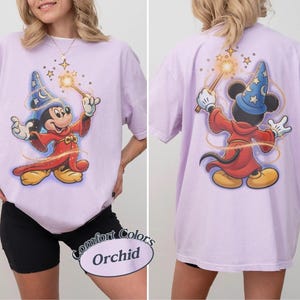May include: An orchid-colored t-shirt featuring a graphic of Mickey Mouse as a sorcerer. The front shows Mickey facing forward with a wand, while the back shows Mickey from behind with a wand. The shirt has short sleeves and a crew neckline.