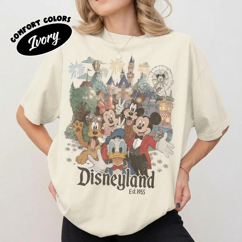 May include: An ivory t-shirt featuring a graphic print of cartoon characters including Mickey Mouse, Minnie Mouse, Donald Duck, Goofy, and Pluto in front of a castle. The text reads Disneyland Est. 1955. The shirt has short sleeves and a crew neckline.