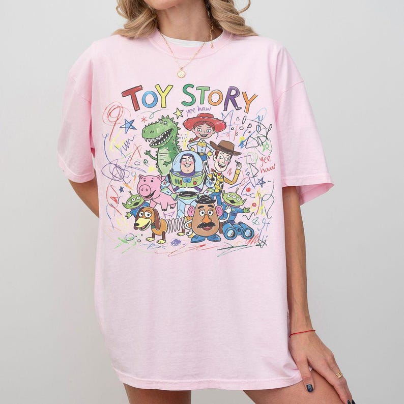 May include: A light pink t-shirt featuring the words "Toy Story" in colorful letters above a graphic of characters from the animated film. The design includes Woody, Buzz Lightyear, and other characters.