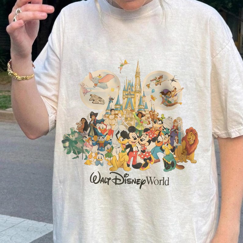 Puede incluir: Camiseta de color crema con un gr&aacute;fico colorido de personajes de Disney, incluyendo Mickey Mouse, Donald Duck y Simba. El dise&ntilde;o incluye el castillo de Cenicienta y el texto "Walt Disney World" en negro.