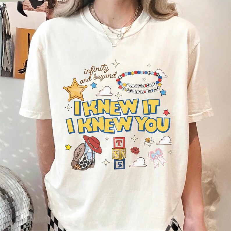 May include: An off-white short-sleeve t-shirt featuring a graphic print with the text "infinity and beyond," "I KNEW IT," "I KNEW YOU," and "YOU'VE GOT A FRIEND IN ME" on a beaded bracelet. Graphics include a star, cowboy boot, blocks, and bow.