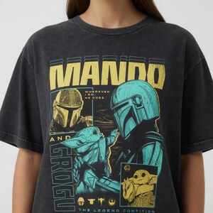 K&ouml;nnte beinhalten: Dunkelgraues T-Shirt mit einem Aufdruck von Mando und Grogu aus The Mandalorian. Das Design enth&auml;lt den Text "MANDO" in Gelb und "GROGU" in T&uuml;rkis, zusammen mit Bildern der Charaktere und dem Satz "Wherever I go, he goes."