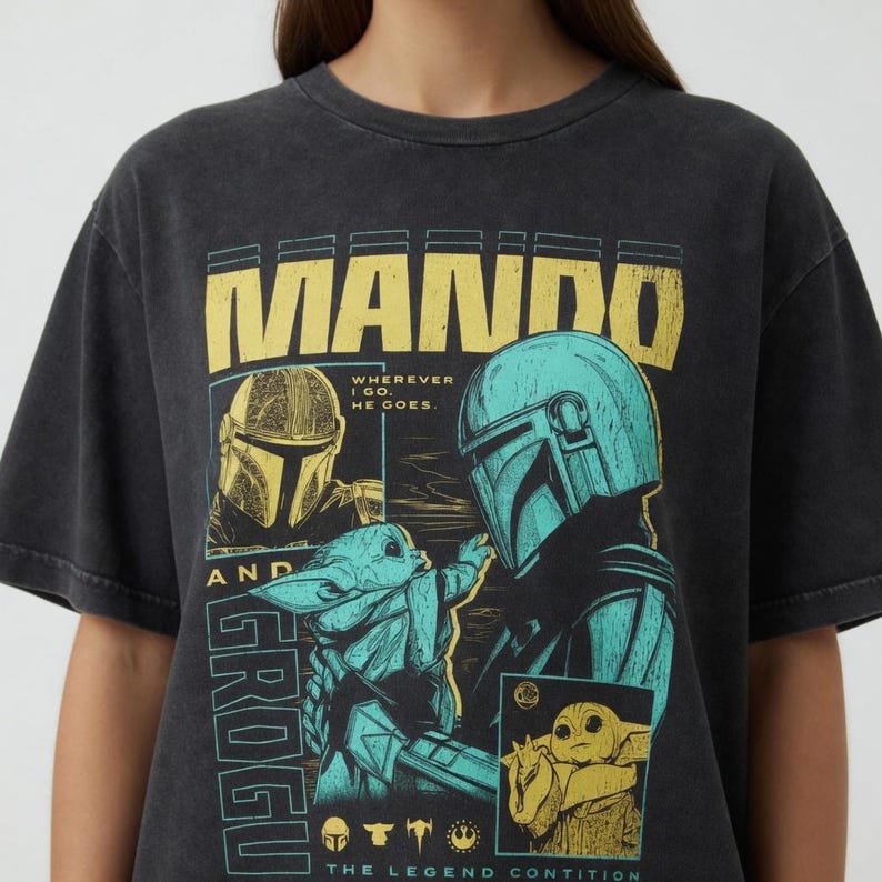 K&ouml;nnte beinhalten: Dunkelgraues T-Shirt mit einem Aufdruck von Mando und Grogu aus The Mandalorian. Das Design enth&auml;lt den Text "MANDO" in Gelb und "GROGU" in T&uuml;rkis, zusammen mit Bildern der Charaktere und dem Satz "Wherever I go, he goes."