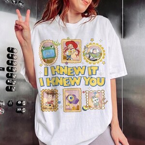 May include: A white short-sleeve t-shirt featuring a graphic print of Toy Story characters in postage stamp frames. The text reads I KNEW IT I KNEW YOU in yellow with blue outlines. Characters include Rex, Jessie, Buzz Lightyear, Woody, and others.