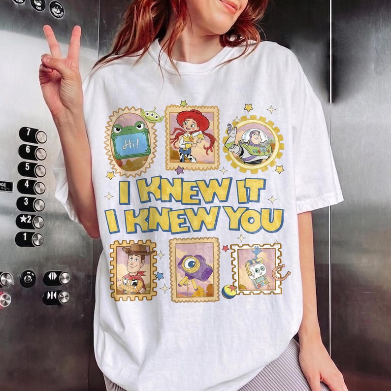 May include: A white short-sleeve t-shirt featuring a graphic print of Toy Story characters in postage stamp frames. The text reads I KNEW IT I KNEW YOU in yellow with blue outlines. Characters include Rex, Jessie, Buzz Lightyear, Woody, and others.