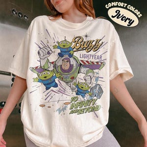Pu&ograve; includere: T-shirt color avorio con una grafica di Buzz Lightyear e dei piccoli alieni verdi di Toy Story. Il design include il testo "Buzz Lightyear" e "To Infinity and Beyond". La maglietta &egrave; etichettata "Comfort Colors Ivory".