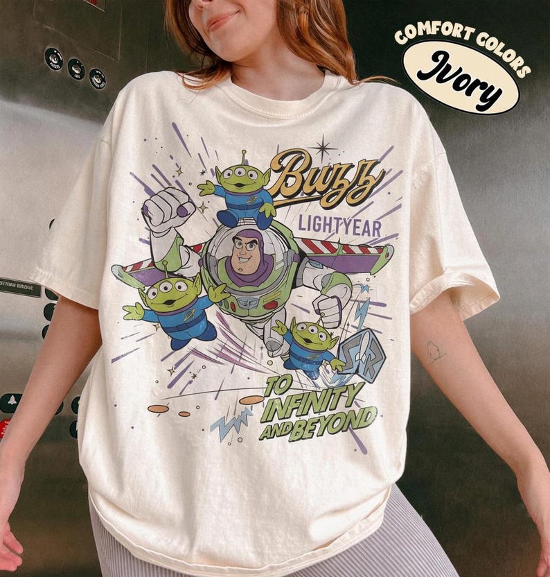 Pu&ograve; includere: T-shirt color avorio con una grafica di Buzz Lightyear e dei piccoli alieni verdi di Toy Story. Il design include il testo "Buzz Lightyear" e "To Infinity and Beyond". La maglietta &egrave; etichettata "Comfort Colors Ivory".