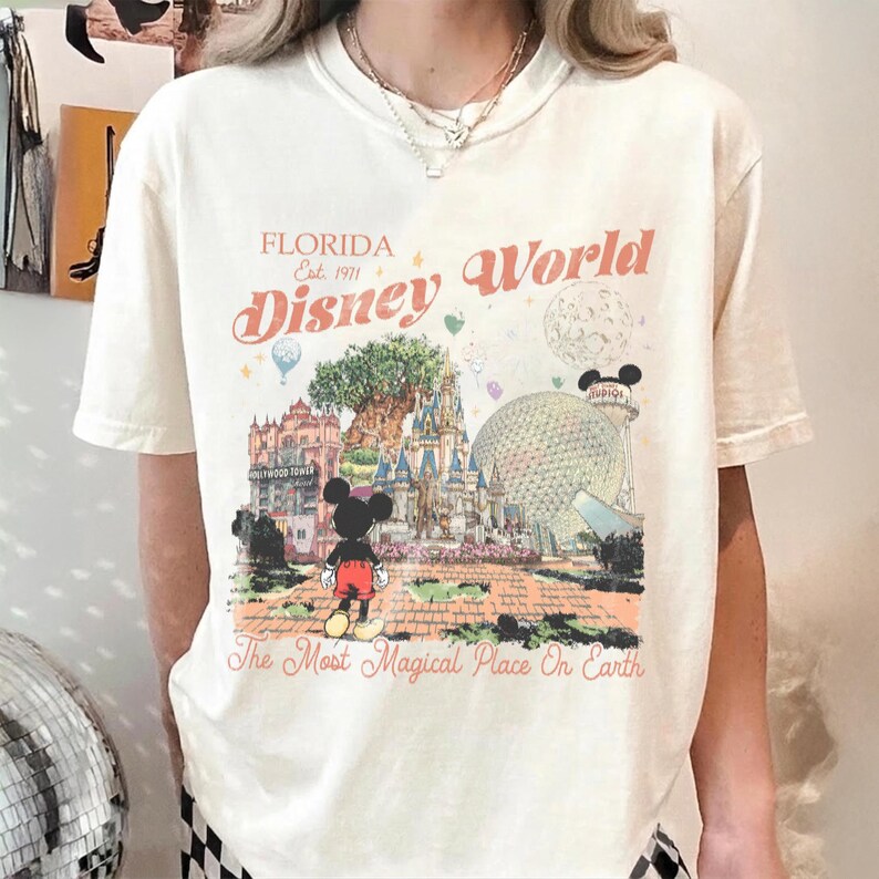 Puede incluir: Camiseta color crema con un gr&aacute;fico colorido de Disney World en Florida. El dise&ntilde;o incluye a Mickey Mouse, un castillo y el texto "Florida Disney World Est. 1971" y "The Most Magical Place On Earth."