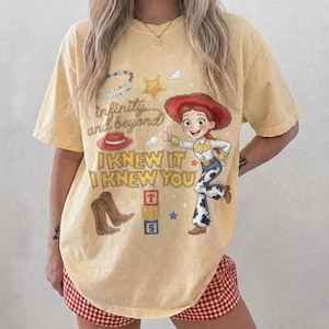 May include: A pale yellow short-sleeve t-shirt featuring a graphic print of a cowgirl character, a red hat, brown cowboy boots, and alphabet blocks. The text reads "infinity and beyond" and "I KNEW IT I KNEW YOU" in a stylized brown font.