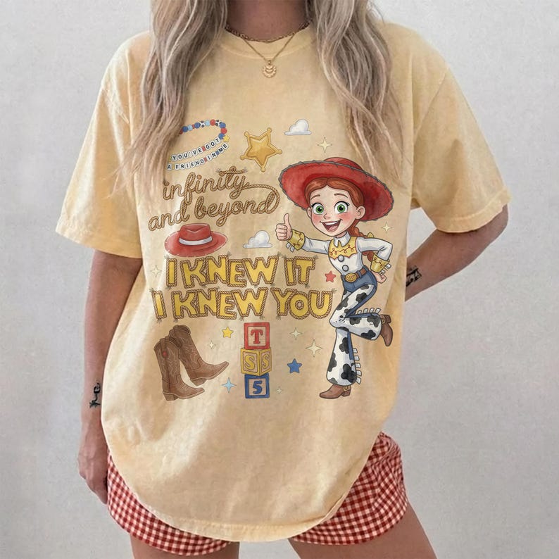 May include: A pale yellow short-sleeve t-shirt featuring a graphic print of a cowgirl character, a red hat, brown cowboy boots, and alphabet blocks. The text reads "infinity and beyond" and "I KNEW IT I KNEW YOU" in a stylized brown font.