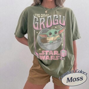 Puede incluir: Camiseta verde oliva con un gr&aacute;fico de Grogu de Star Wars. El dise&ntilde;o incluye el texto "THE CHILD" y "GROGU" en rosa, junto con el logotipo de Star Wars. La camiseta est&aacute; etiquetada como "Comfort Colors Moss".
