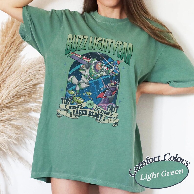 May include: A sage green short-sleeve t-shirt featuring a graphic of Buzz Lightyear from Toy Story. The design includes the text BUZZ LIGHTYEAR, DISNEY TOY STORY, and LASER BLAST, with illustrations of Buzz Lightyear, Zurg, and green aliens in space.