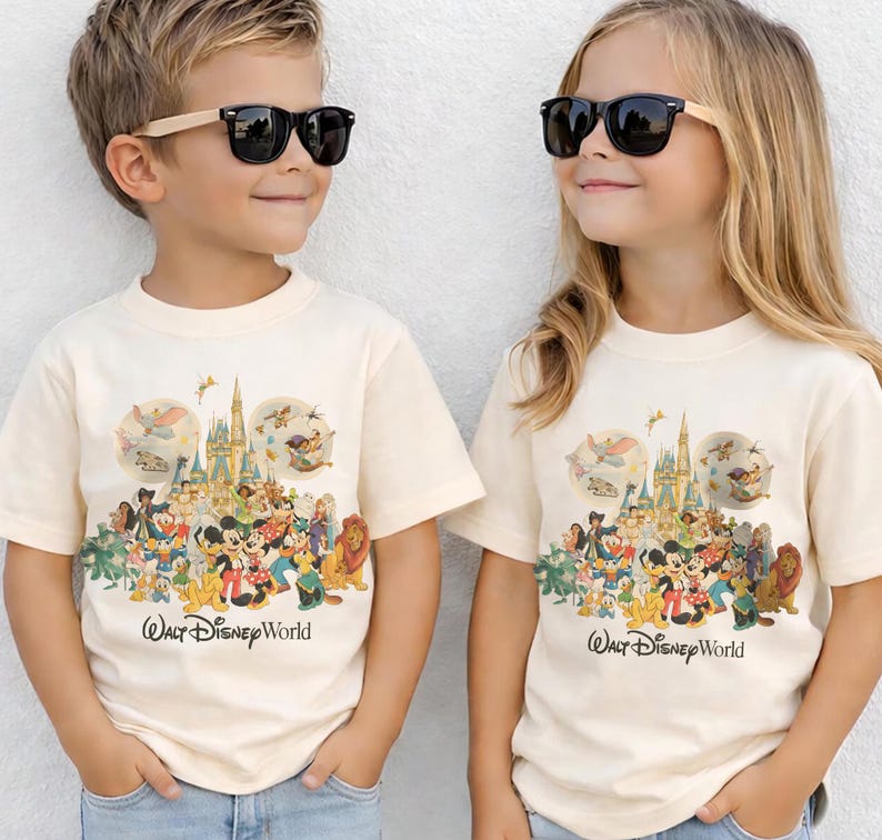 Puede incluir: Dos ni&ntilde;os vistiendo camisetas color crema con un gr&aacute;fico colorido de personajes de Disney y un castillo. Las camisetas tienen el texto "Walt Disney World" impreso. Ambos ni&ntilde;os llevan gafas de sol negras.