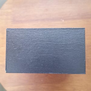 May include: A rectangular, black wooden box with a textured surface. The box is positioned on a round, wooden table. The box appears to be empty.