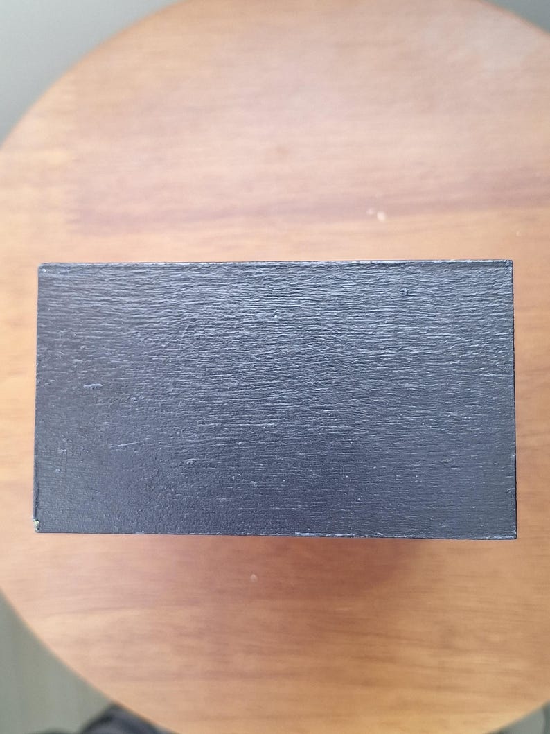 May include: A rectangular, black wooden box with a textured surface. The box is positioned on a round, wooden table. The box appears to be empty.