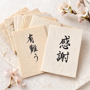 以下が含まれることがあります： 黒インクの日本語カリグラフィーが施された、小さな長方形のカードの束。1枚のカードには「ありがとう」、もう1枚には「感謝」と書かれています。カードは薄いベージュ色で、桜の花が咲く白い表面に配置されています。
