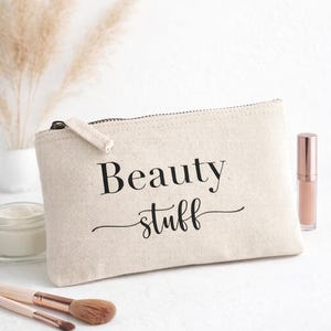 Könnte beinhalten: Eine beige Kosmetiktasche aus Canvas mit dem Schriftzug "Beauty stuff" in schwarzer Schrift. Die Tasche hat einen Reißverschluss und liegt auf einer weißen Oberfläche, mit Make-up-Pinseln und einer Tube Lipgloss in der Nähe. Im Hintergrund eine Vase mit Trockenblumen.